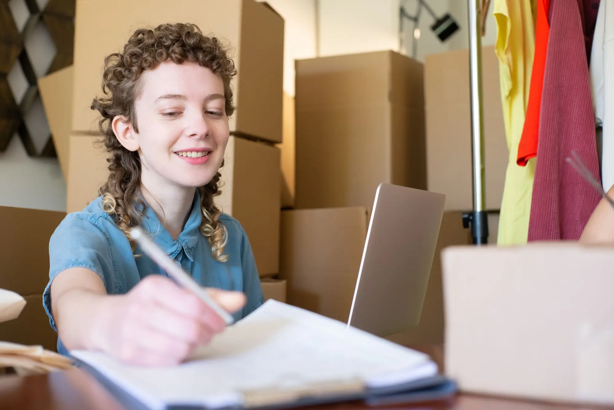 Small business owner in her stockroom ticking off customer invoices on a clipboard beside her laptop, surrounded by boxed orders and a clothing rack