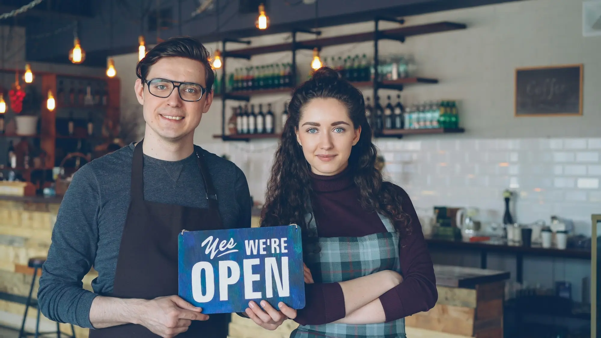 Two young café co-owners in aprons holding a 'Yes we're open' sign at the counter of their new coffee shop