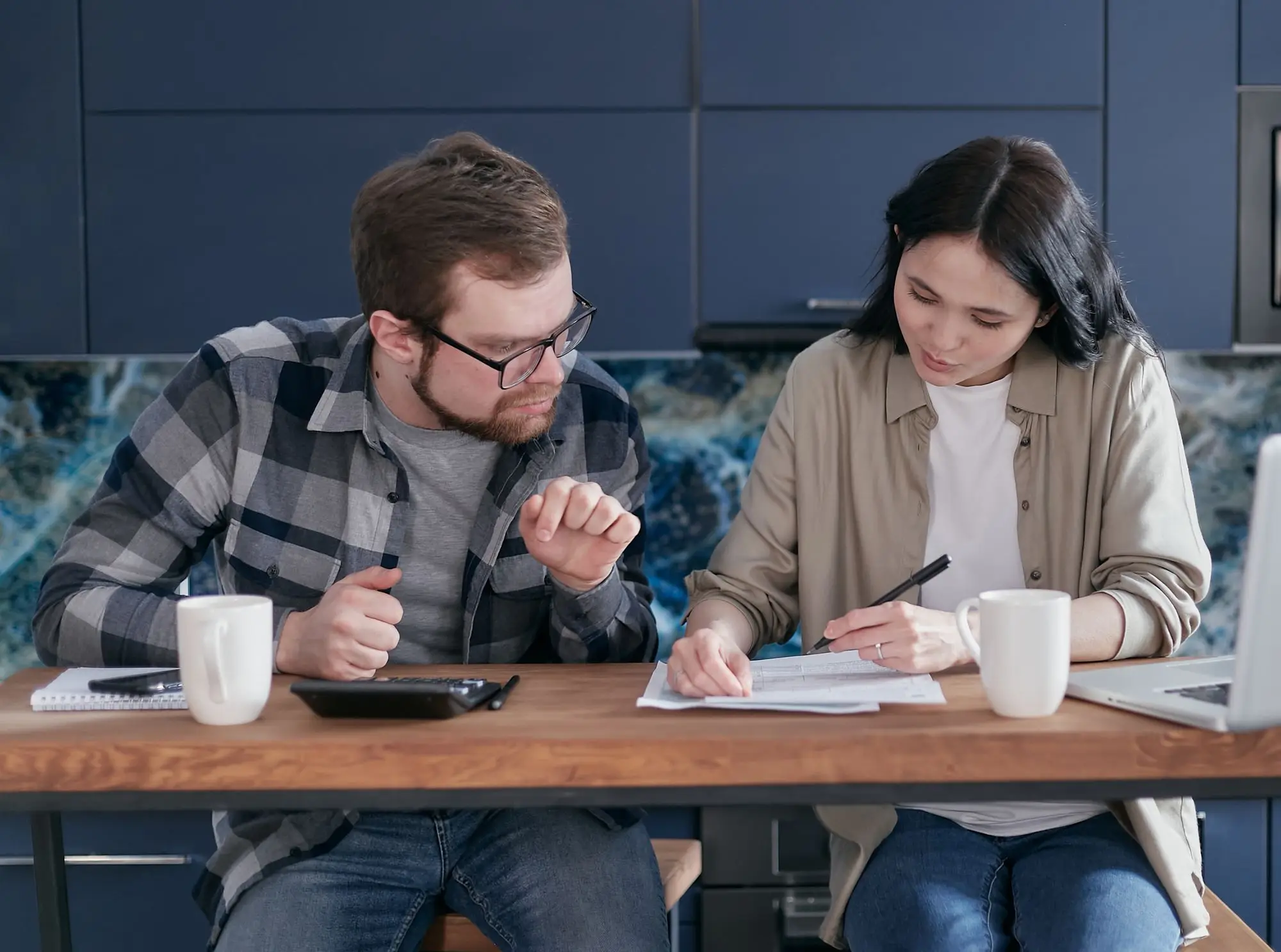 Business owner and partner at a kitchen bench with a calculator, laptop and home loan paperwork, weighing up a redraw for the business